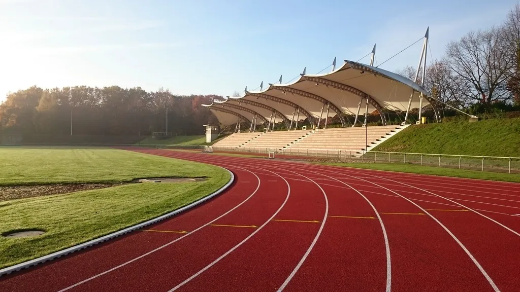 Pista de atletismo de pavimento sintético rojo al amanecer, con una grada cubierta al fondo y la hierba del interior cubierta de rocío