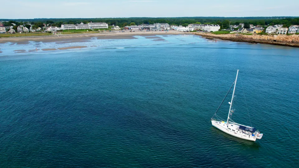 Velero de casco blanco navegando solo en una bahía de aguas azules con la costa al fondo
