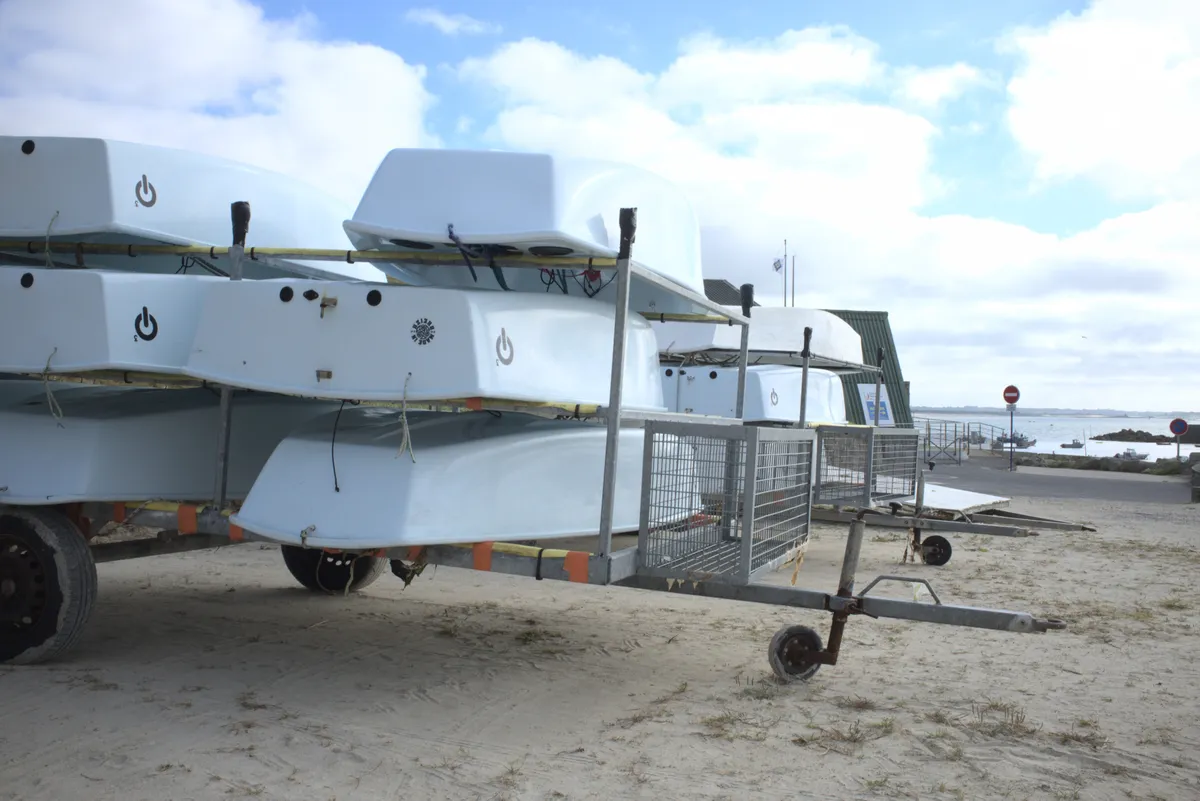 Embarcaciones ligeras de vela apoyadas sobre remolques en la arena de una playa, con el mar y el cielo nublado al fondo